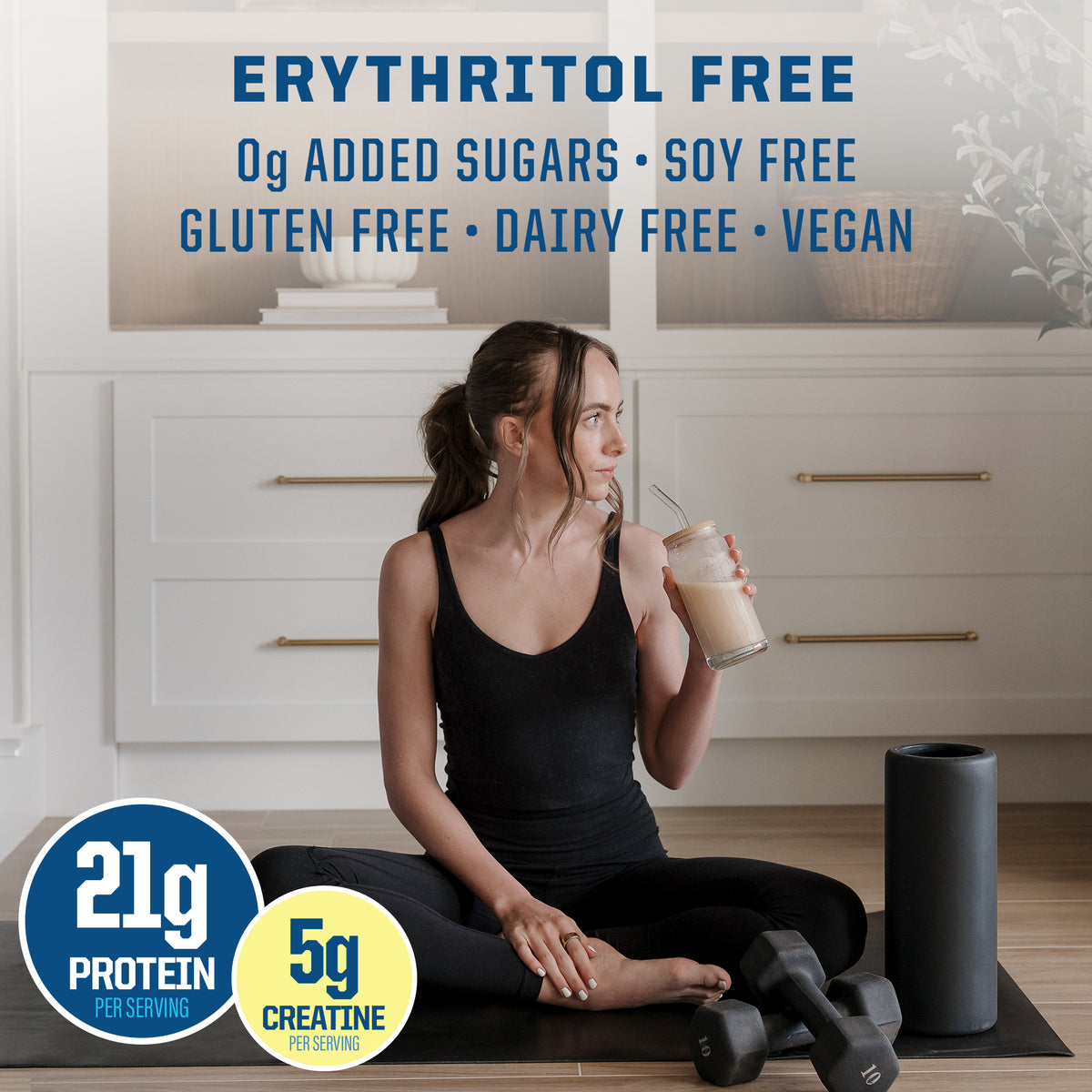Woman in black workout clothes sitting on mat drinking shake near weights and foam roller in bright room
