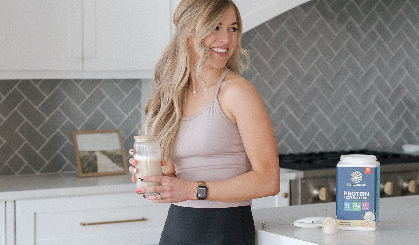 A woman in a kitchen holding a protein shake, with a blue tub of Sunwarrior Protein Creatine Powder on the counter next to her.